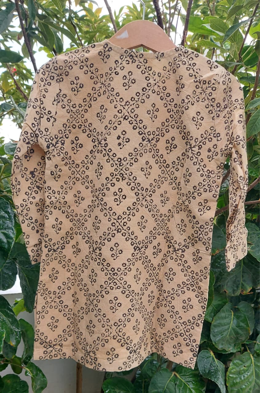 A beige long-sleeve tunic or kurta with a black geometric block print pattern, hanging on a wooden hanger against a background of green leaves.