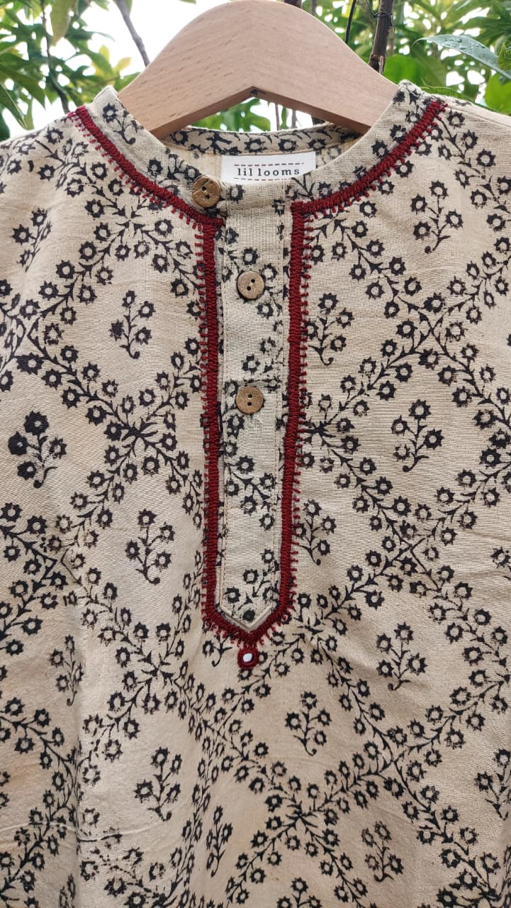 A close-up of a cream-colored cotton tunic top with a black floral block print, featuring a red embroidered border along the split neckline and button placket. The top is displayed on a wooden hanger against a background of green foliage.