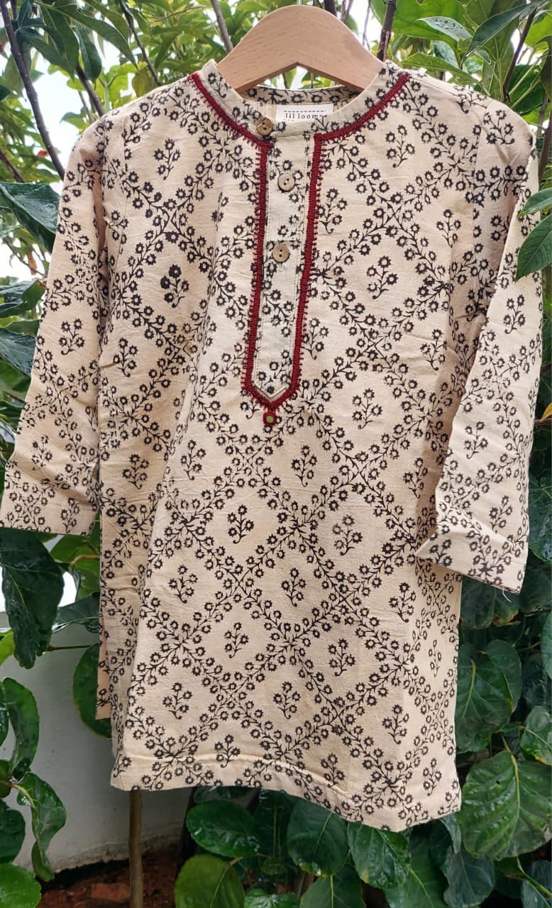 A beige cotton tunic top with a black floral block print and red embroidered trim along the neckline, hanging on a wooden hanger against a background of green foliage.
