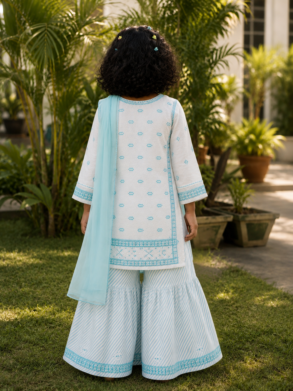A child viewed from behind wearing a light blue and white traditional outfit with matching wide-leg pants and a sheer dupatta, standing outdoors on grass with potted plants in the background.