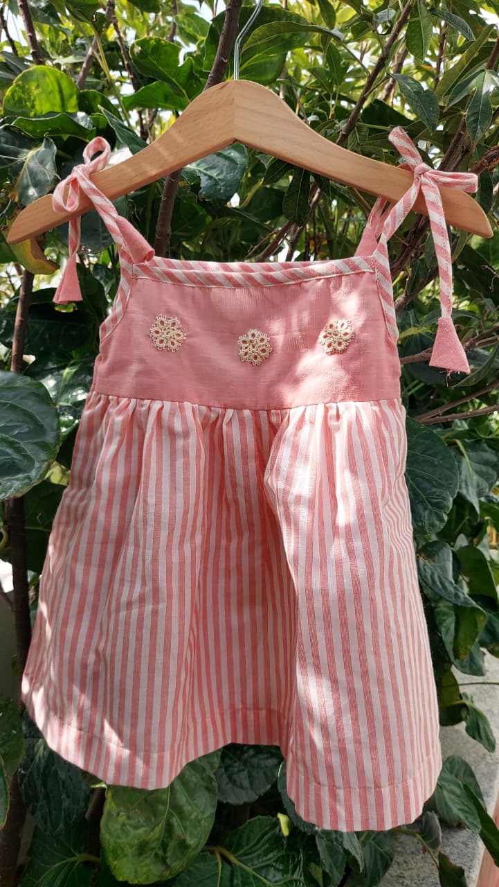 A pink children's sleeveless romper with vertical pink and white stripes, featuring a solid pink yoke decorated with three small floral appliqués and tied shoulder straps, hanging on a wooden hanger against a background of green garden foliage.