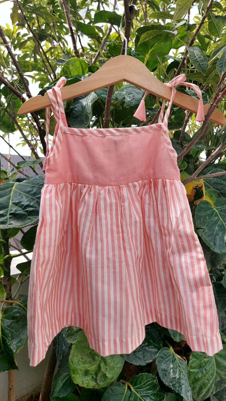 A pink sleeveless children's dress with thin shoulder ties and vertical pink-and-white stripes, hanging on a wooden hanger outdoors in front of green leaves.