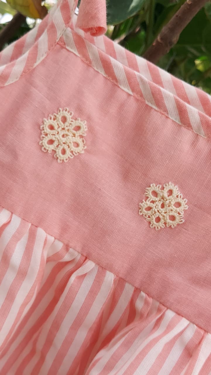 A close-up of a pink and white striped fabric garment, likely a dress or apron, featuring two cream-colored floral lace appliques on the front panel.