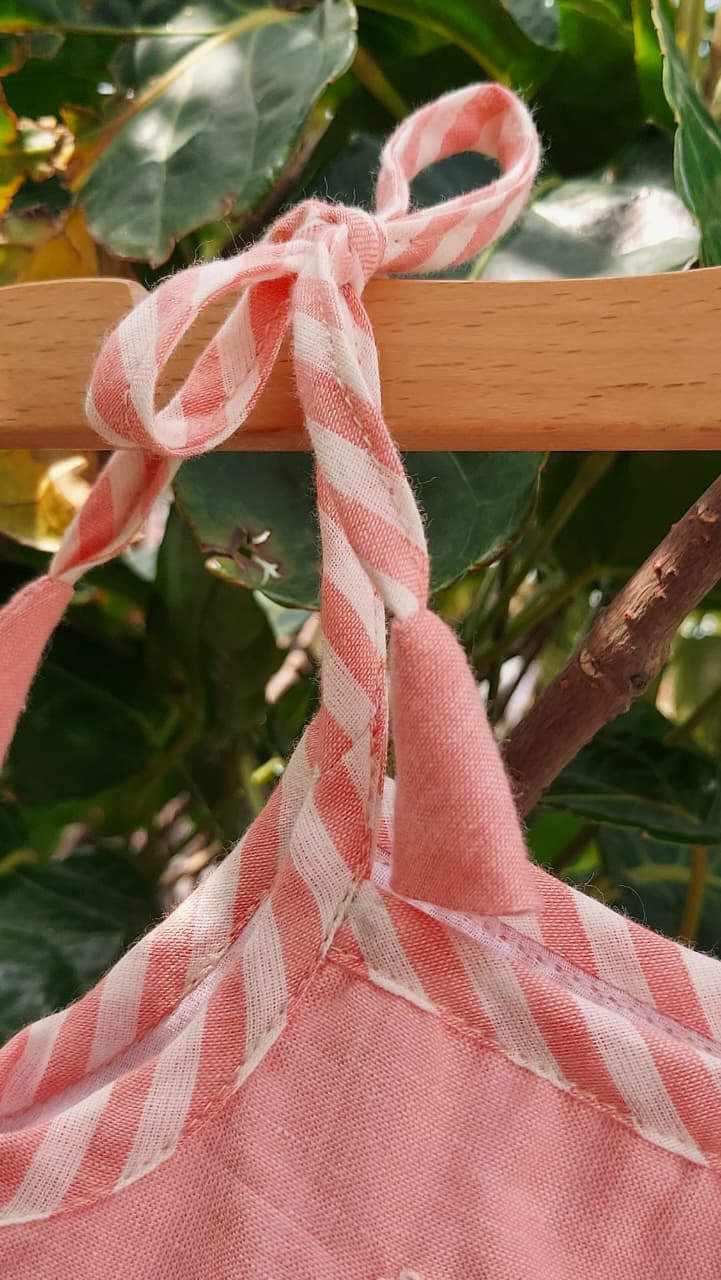 Pink and white striped fabric garment hanging from a wooden hanger outdoors, with green leaves and branches in the background.