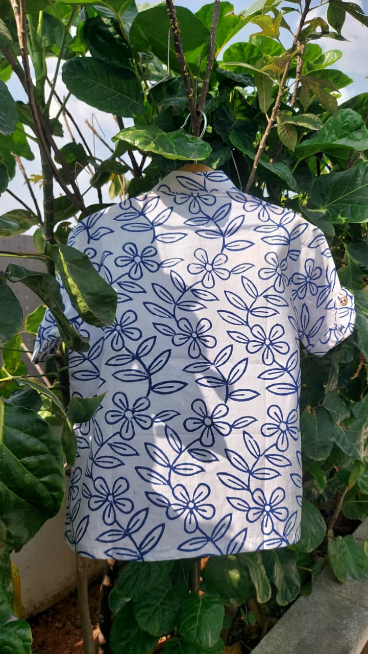 A short-sleeve button-up shirt with a white base and a dark blue floral line pattern, hanging outdoors among green leaves.