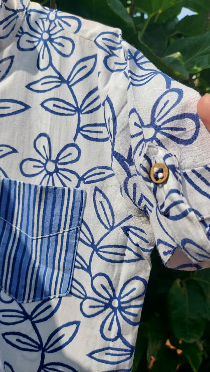 A close-up of a short-sleeve button-up shirt with a blue floral block print on an off-white base, featuring a solid blue striped pocket and a brown button.