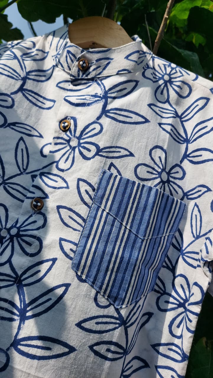 A close-up of a white cotton shirt with a blue floral block print pattern and a contrasting blue-and-white striped pocket, hanging on a wooden hanger against a background of green leaves.