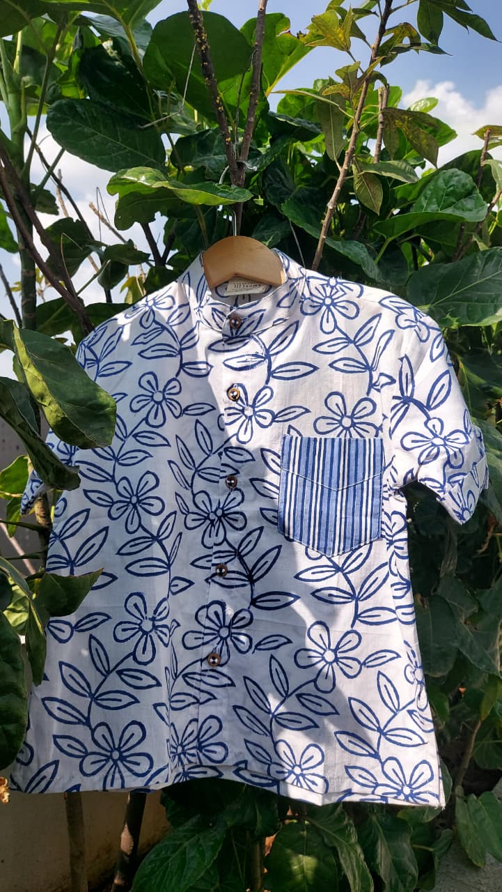 A white short-sleeve button-up shirt with a blue floral line pattern and a blue-and-white striped chest pocket, hanging on a wooden hanger outdoors among green leaves.