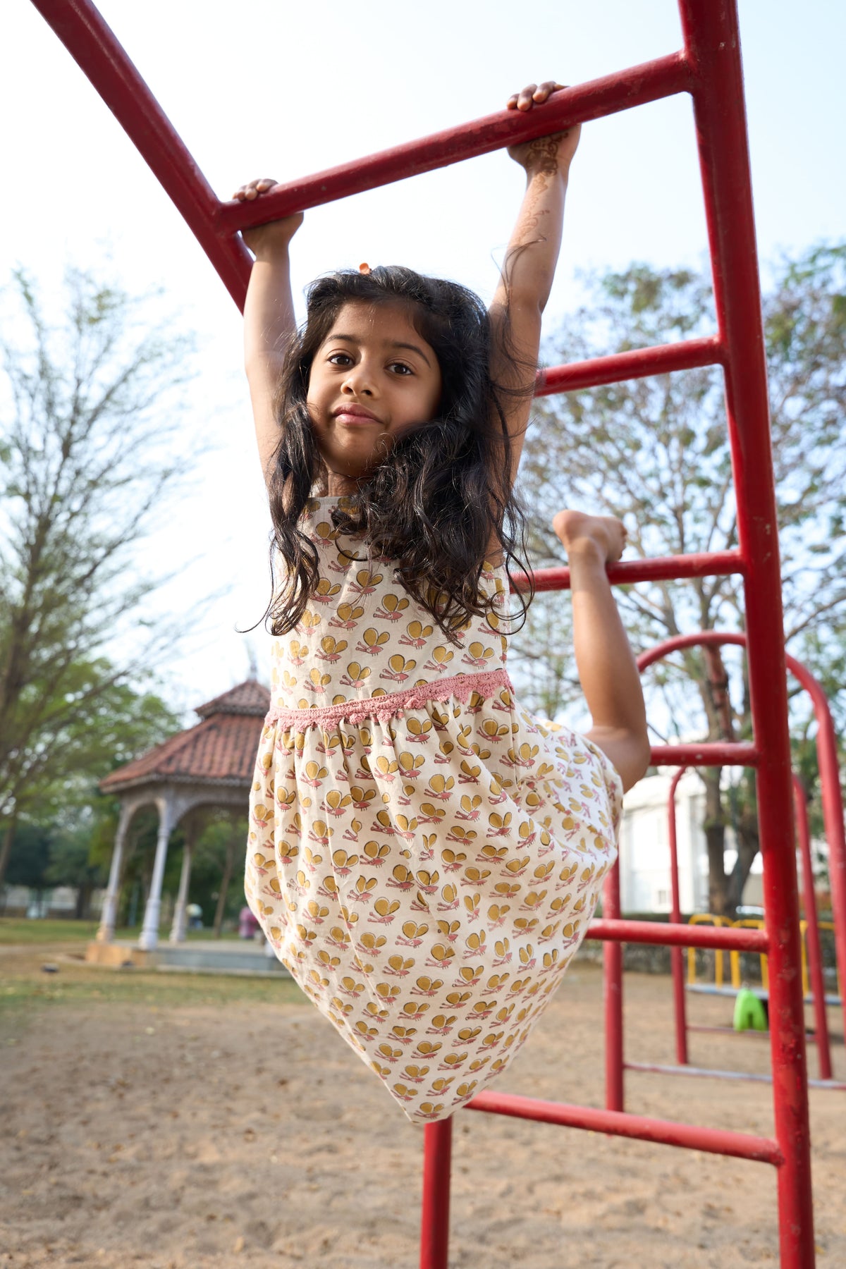 Young girl in a patterned dress hanging from a red playground structure.