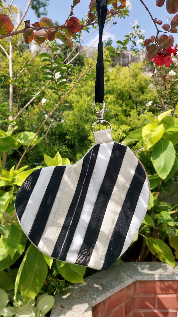 A heart-shaped black and white striped fabric bag with a black hanging strap, photographed outdoors against a background of green foliage and a brick wall.
