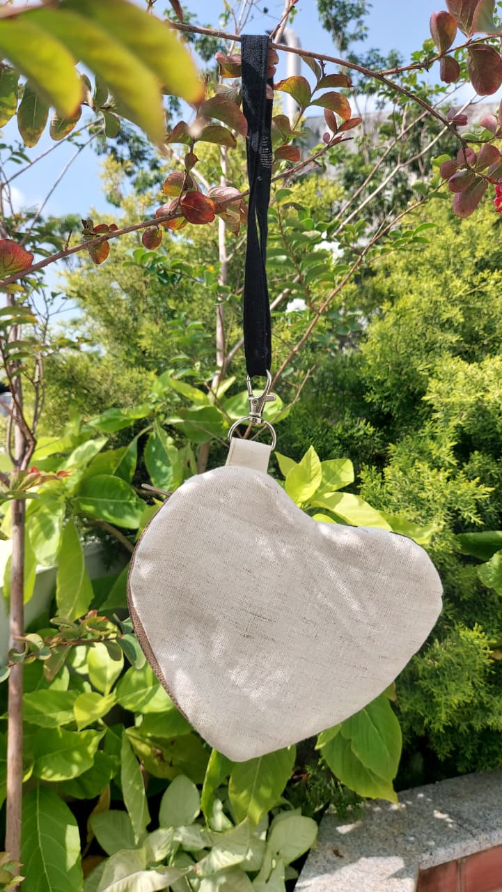 A heart-shaped white wooden hanging ornament with a black ribbon loop, suspended from a tree branch outdoors against a background of green leaves and blue sky.