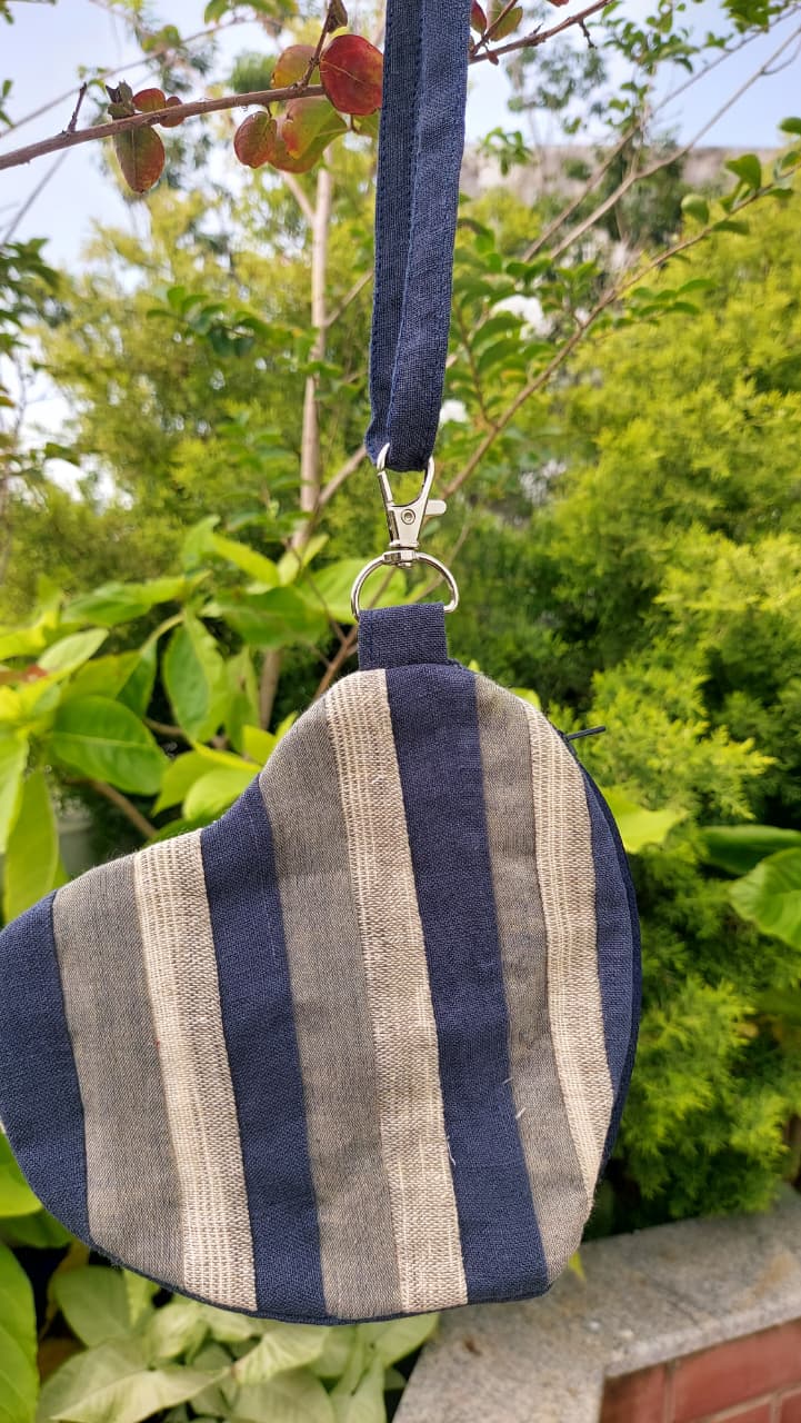 A small fabric shoulder bag with vertical navy blue and beige stripes, hanging from a metal ring on a tree branch outdoors with green foliage in the background.