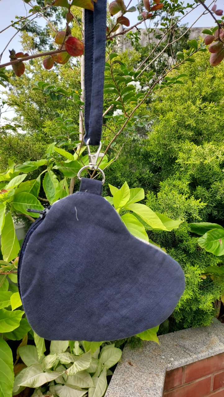 A dark navy blue heart-shaped shoulder bag made of textured fabric, hanging from a matching strap with a metal clasp against a background of green garden foliage.