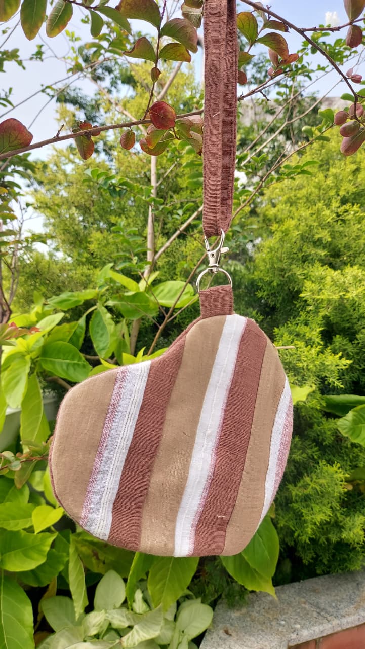 A small heart-shaped fabric pouch with vertical brown, white, and beige stripes, hanging from a brown strap outdoors among green leaves and branches.