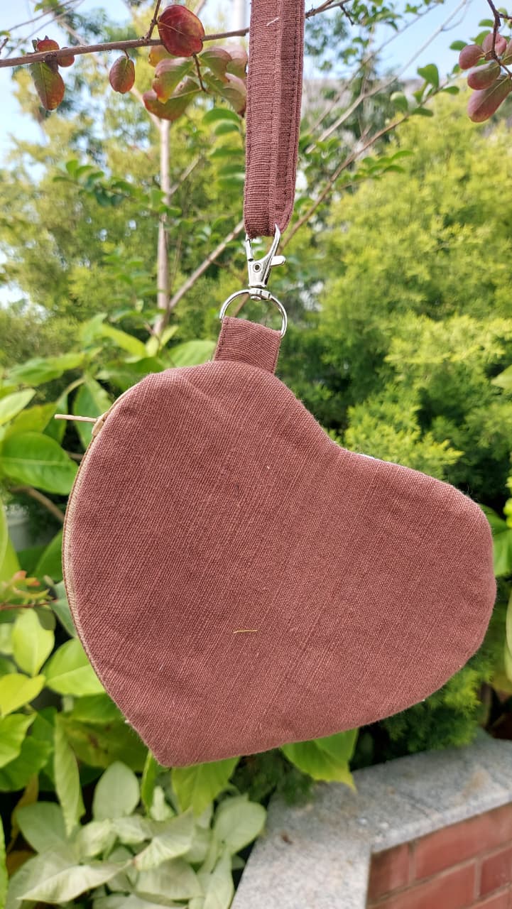 A heart-shaped dusty rose fabric pouch with a matching wrist strap and silver metal clasp, hanging outdoors against a background of green foliage and small red flowers.