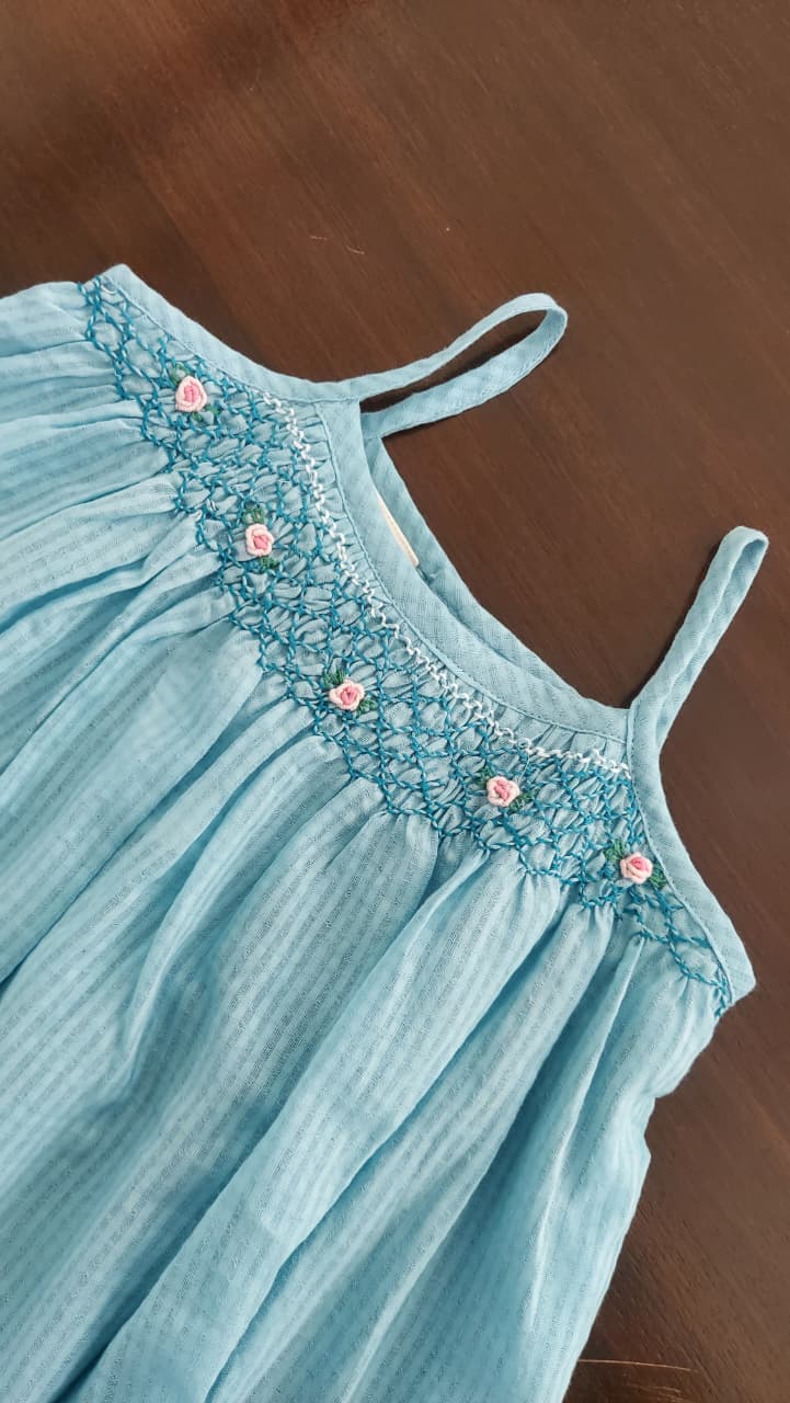 A light blue sleeveless smocked dress with delicate lace embroidery and small pink flower accents along the neckline, laid flat on a dark brown wooden surface.