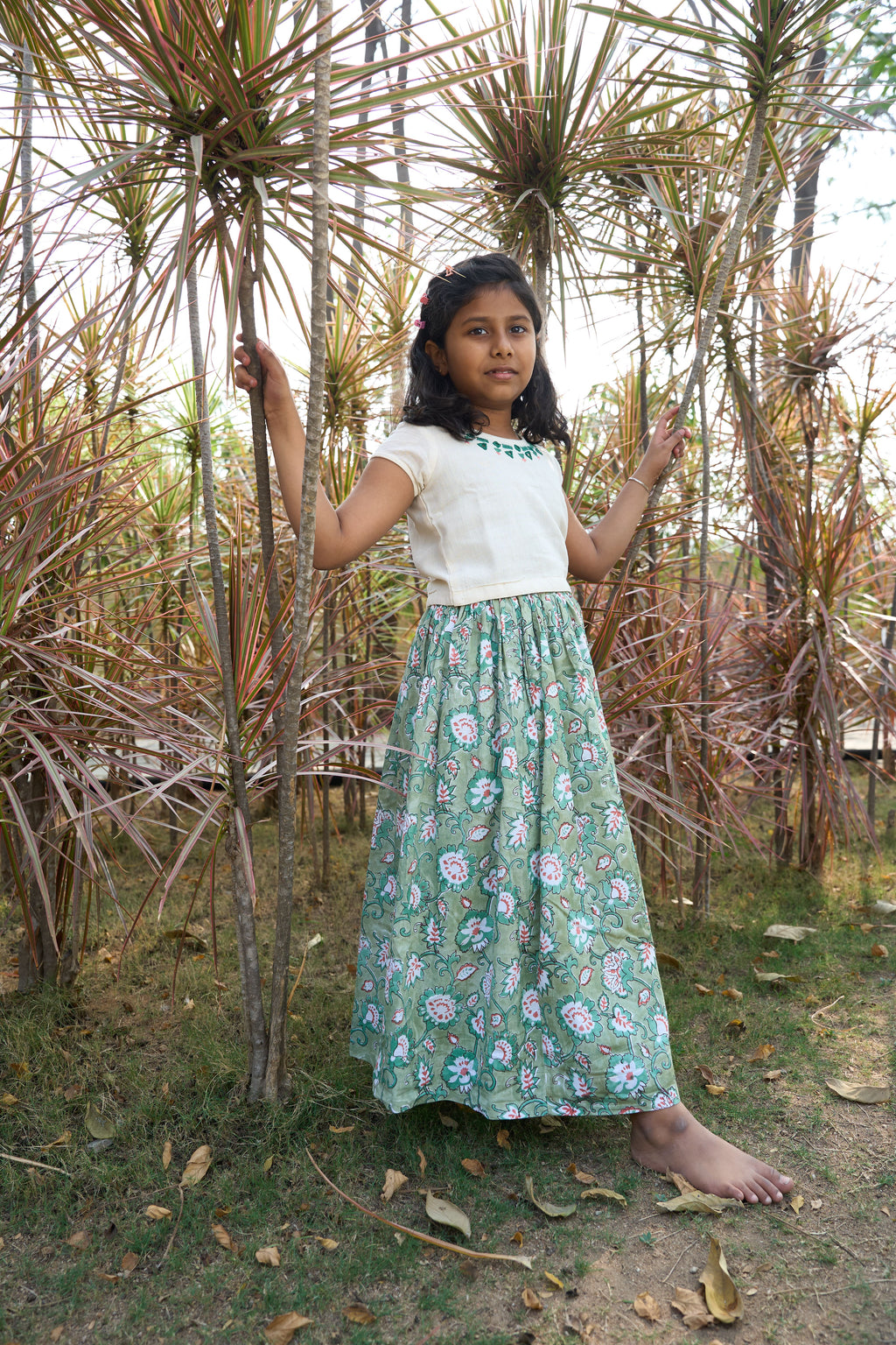 Young girl in a white top and green floral skirt standing among dried plants.