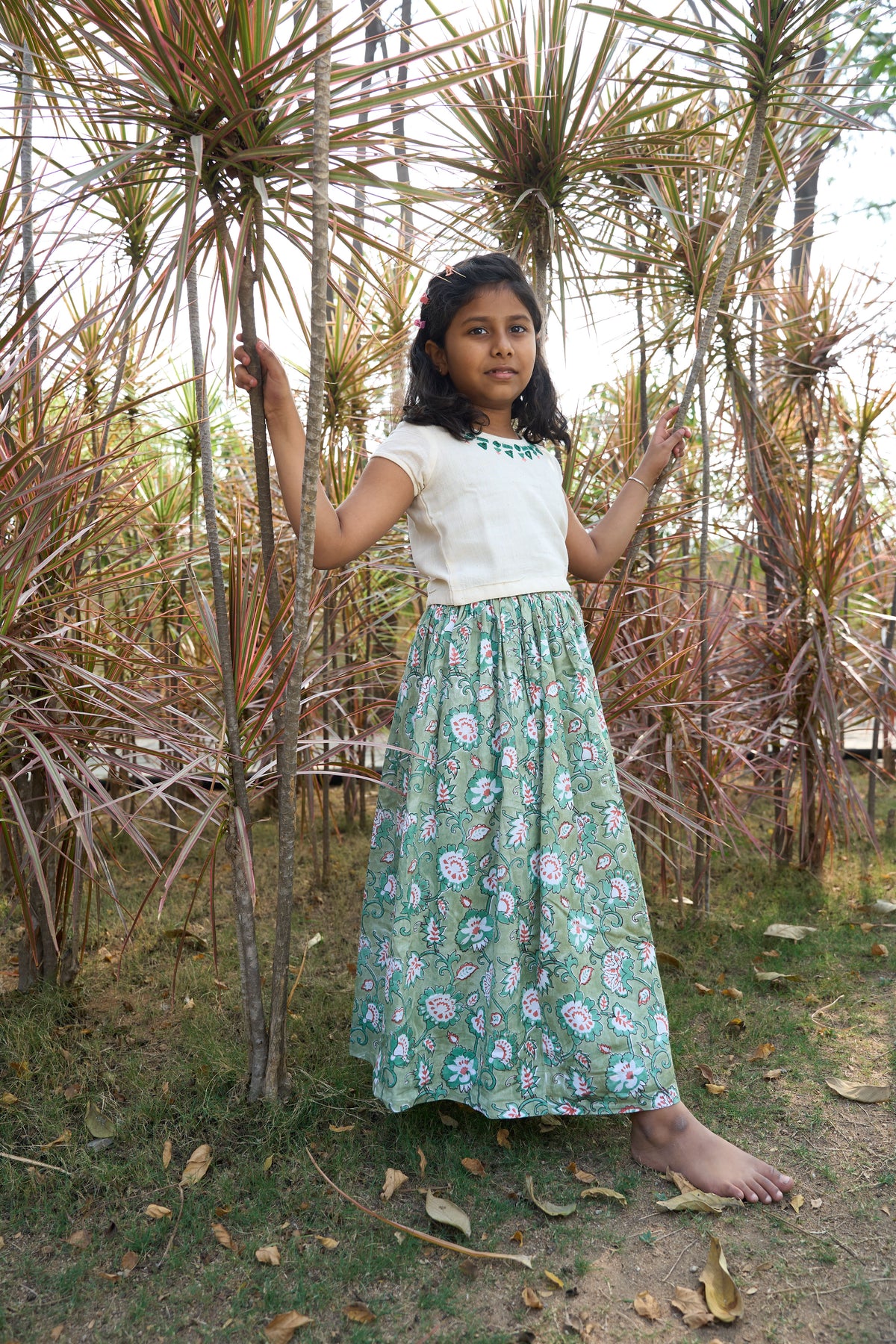 Young girl in a white top and green floral skirt standing among dried plants.