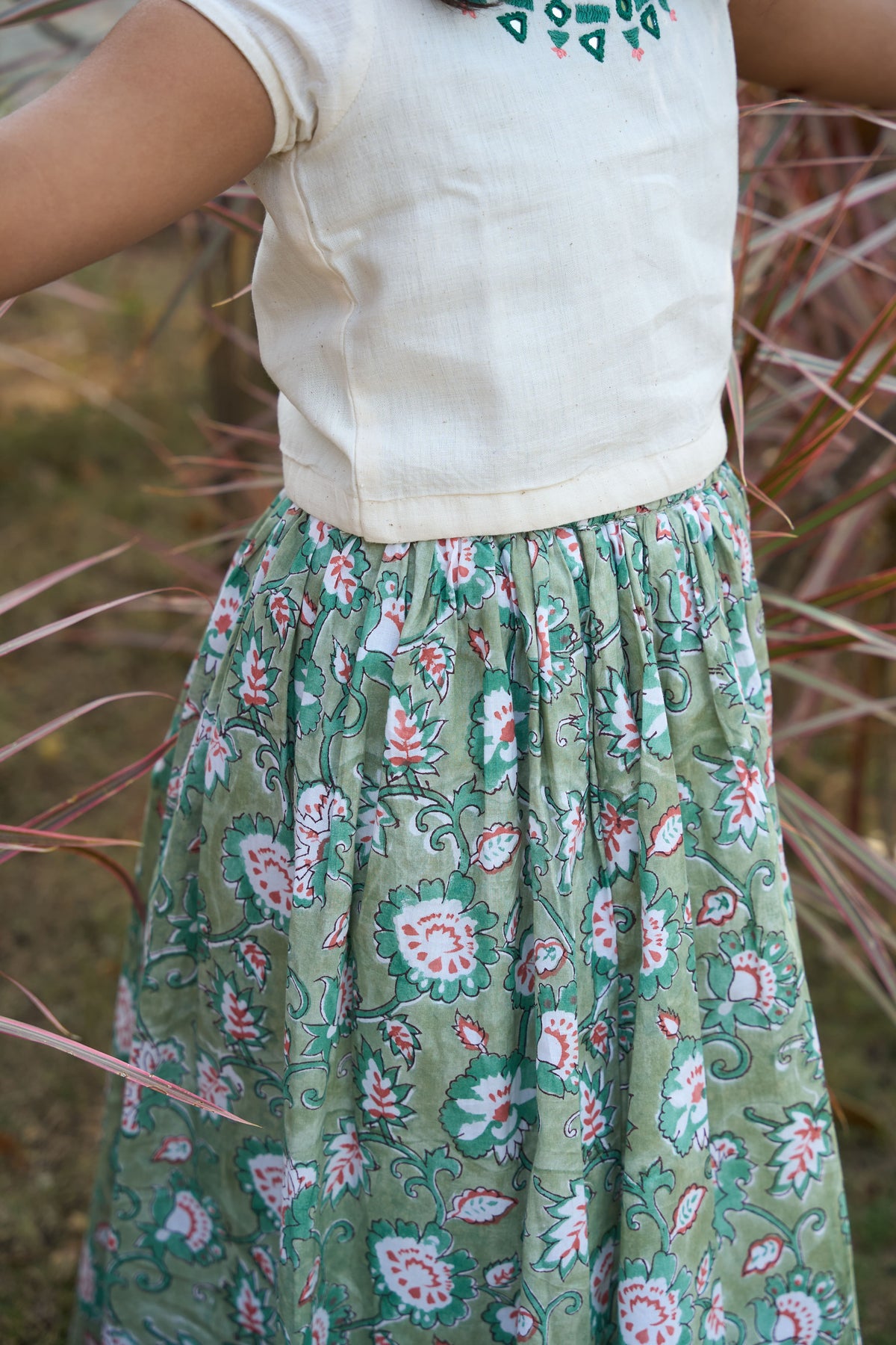 Person wearing a white top and green floral skirt standing in a natural setting.