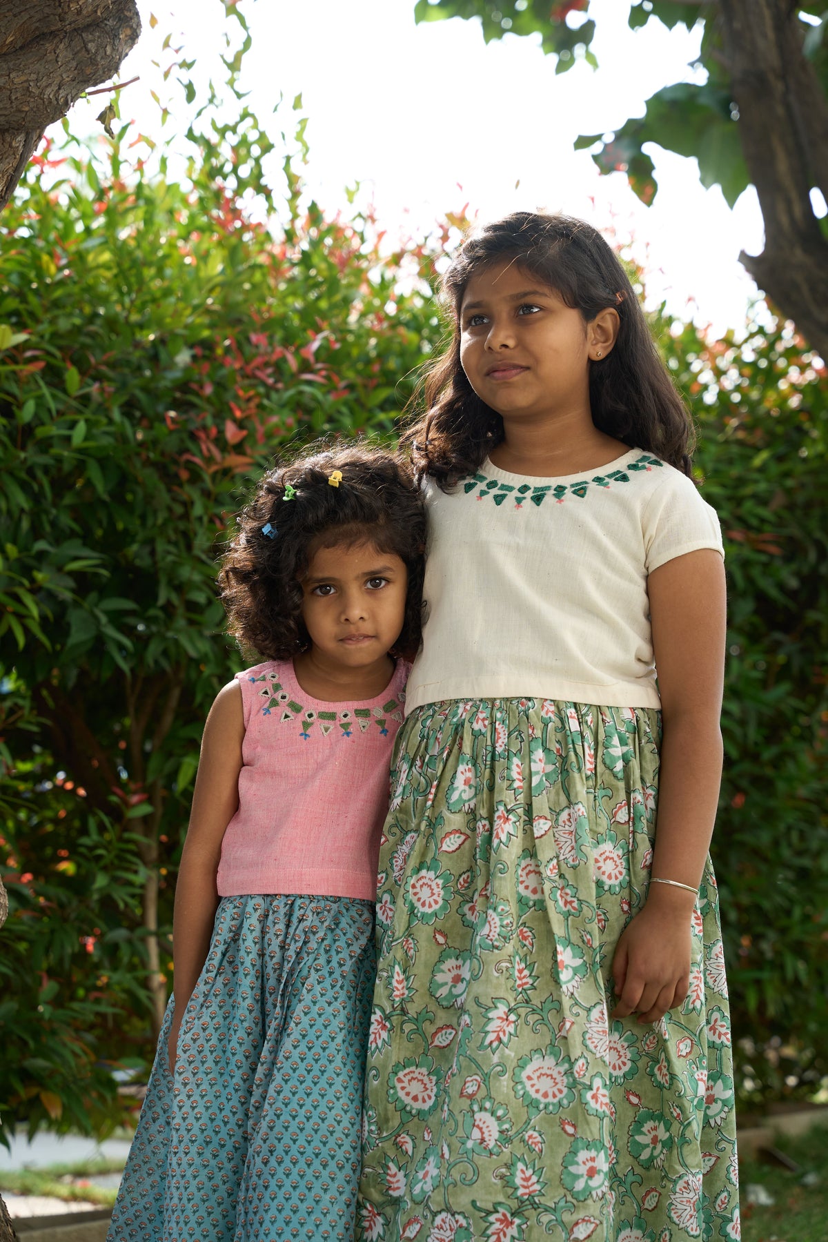 Two young girls standing together outdoors with greenery in the background