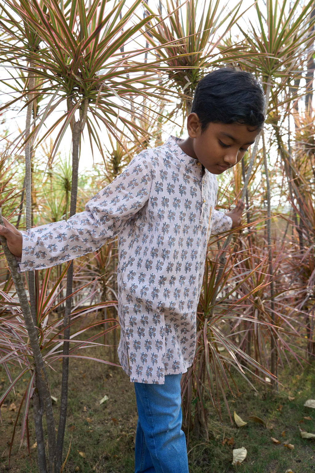 Child in a patterned shirt and blue pants standing among tall plants