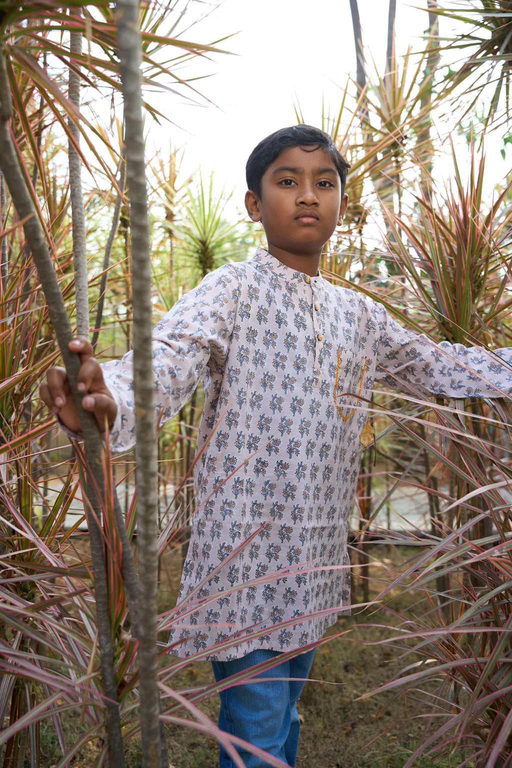 Person wearing a long, light-colored floral shirt standing among tall plants.