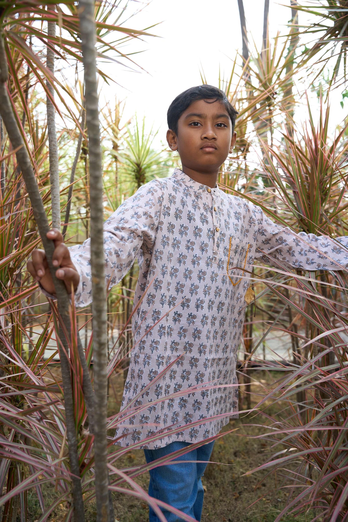 Person wearing a long, light-colored floral shirt standing among tall plants.