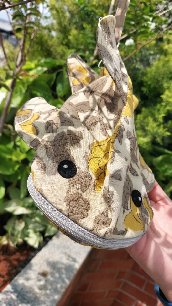 AFish-shaped zippered pouch with a beige, brown, and yellow floral pattern, held by a hand outdoors against a background of green foliage.
