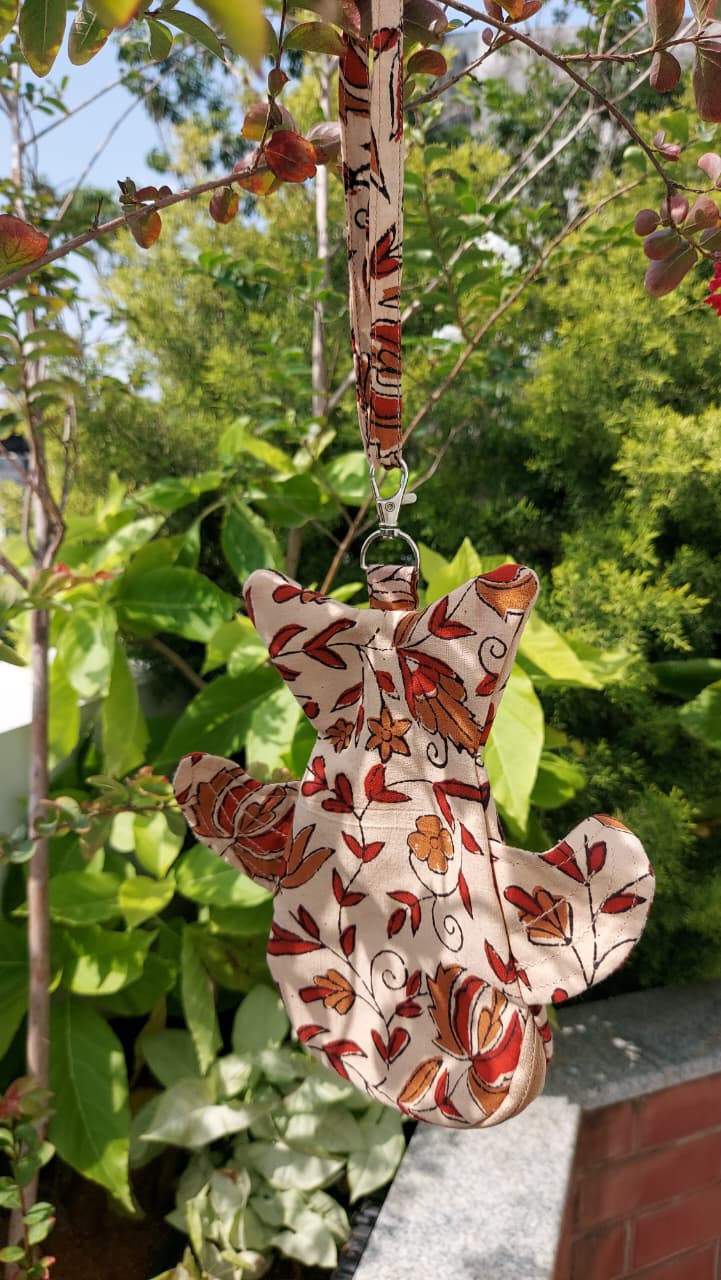 A fabric hanging ornament shaped like a fox, featuring a red and orange autumn leaf pattern on a cream background, suspended by a matching patterned strap outdoors against a backdrop of green garden foliage.