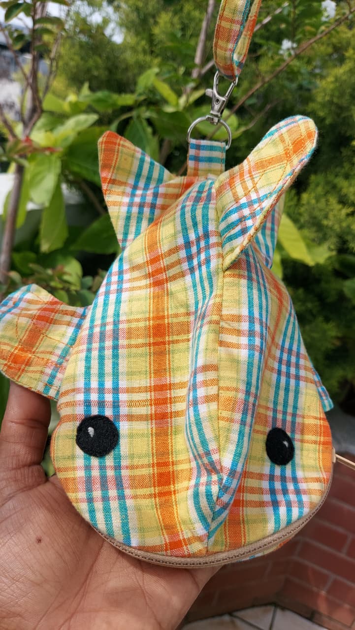 A small plaid pouch shaped like a fox or rabbit head, held in a hand outdoors. The fabric has a yellow, orange, green, and blue plaid pattern with black embroidered eyes and a matching wrist strap at the top.