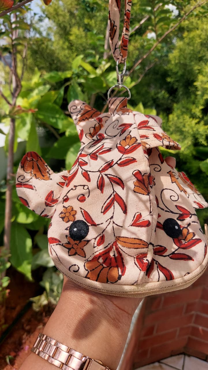 A hand holding a small fabric pouch or bag with a floral print, featuring orange and red flowers on a beige background, with a metal clasp and strap at the top. The background shows green foliage and a brick wall.
