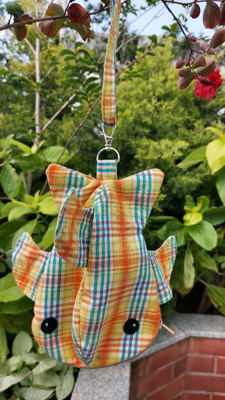 A handmade fish-shaped fabric bag with a plaid pattern in orange, green, and yellow, featuring black bead eyes and a matching strap, hanging outdoors against a background of green foliage.