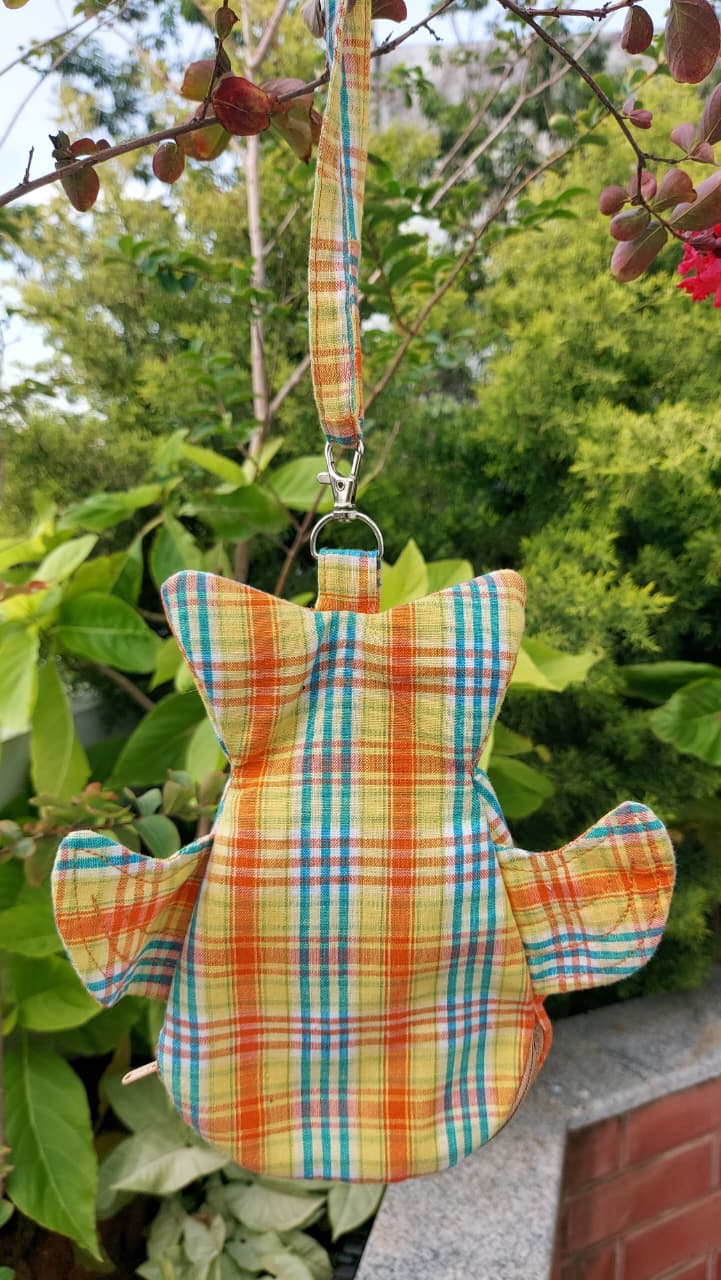 A small plaid fabric accessory with a strap and flap details, hanging outdoors against a background of green leaves and branches. The fabric features a multicolor plaid pattern in orange, yellow, blue, and green.