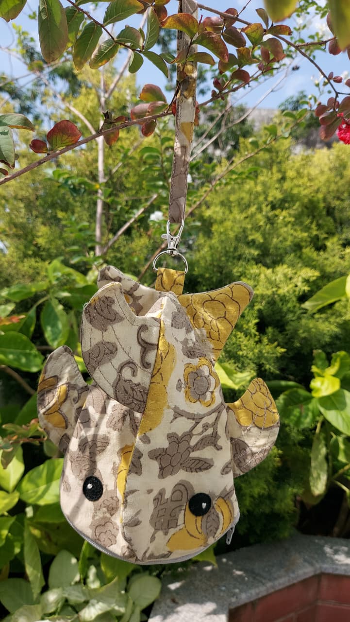 A small fabric animal-shaped pouch or bag with a yellow and brown pattern, hanging from a metal clip attached to a tree branch outdoors in front of green foliage.