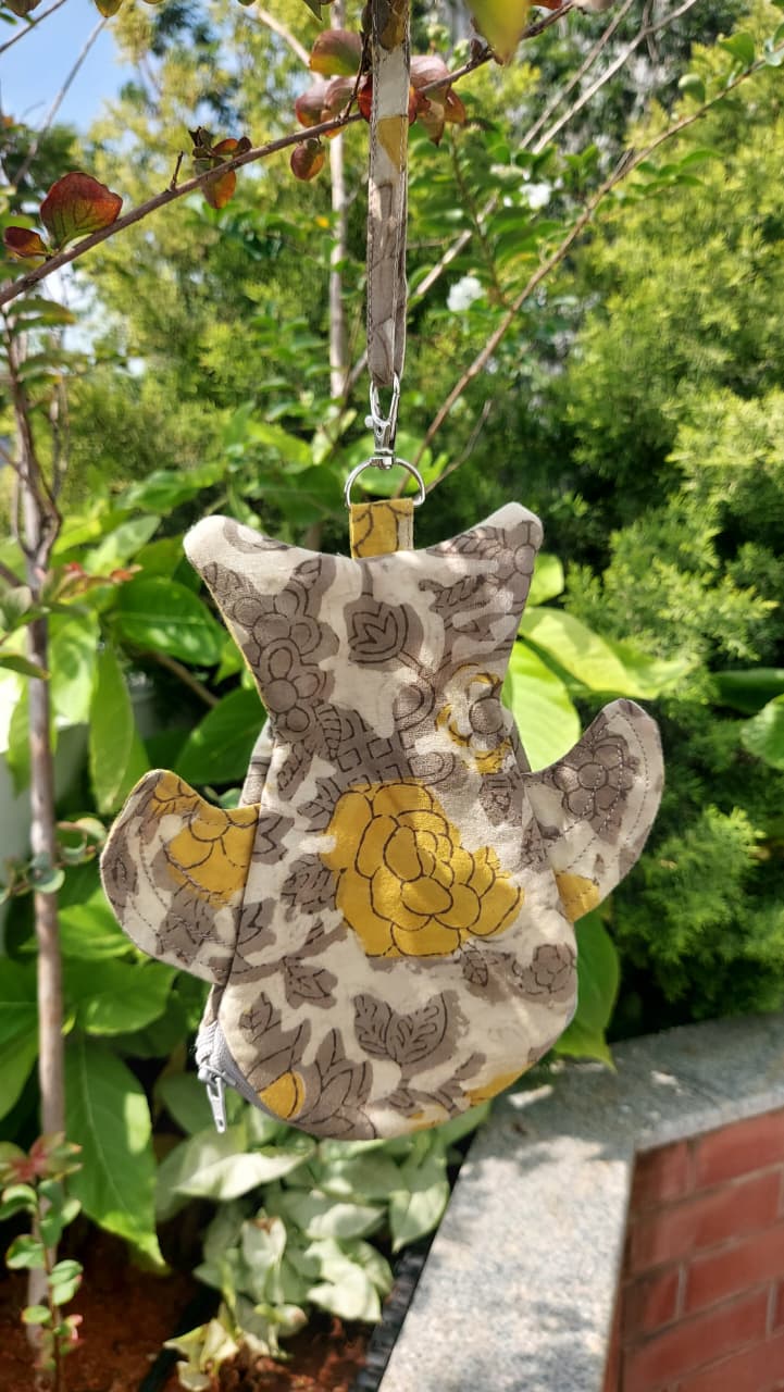 A small fabric bird-shaped pouch or organizer hanging from a tree branch outdoors, featuring a floral pattern in yellow, brown, and beige against a green garden background.