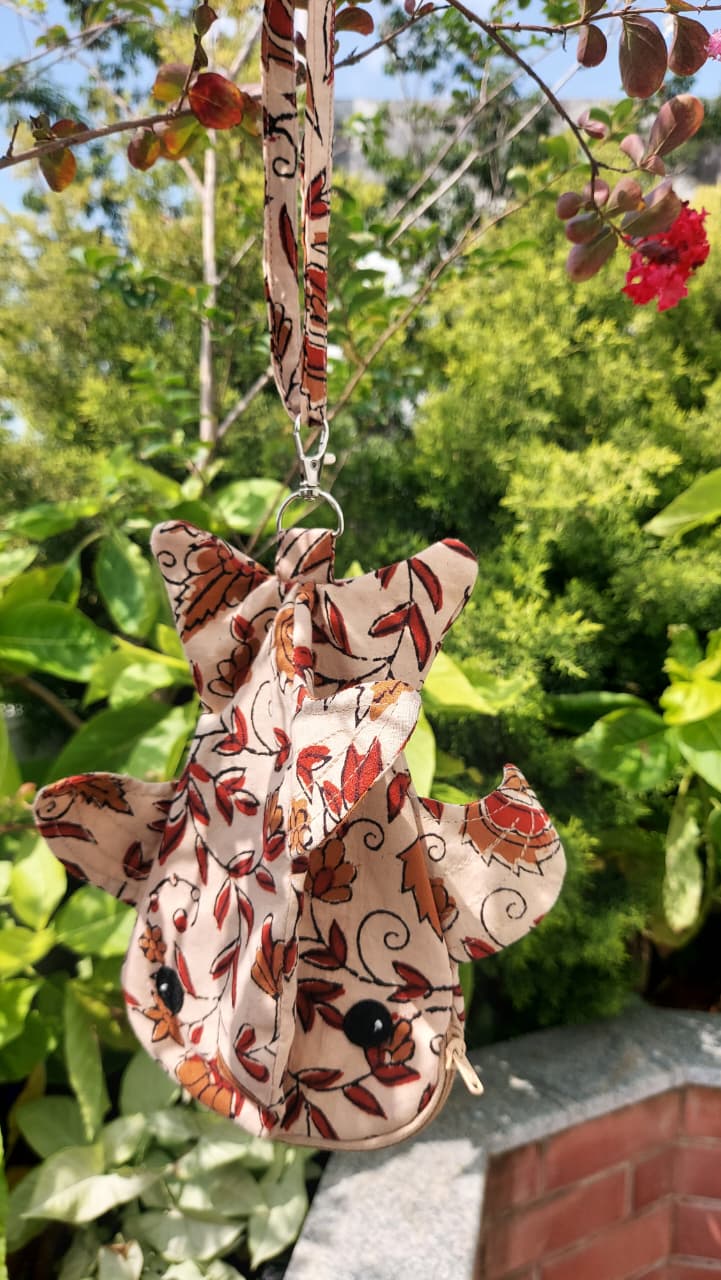 A small fabric animal-shaped pouch with a butterfly design, featuring a brown and beige floral print, hanging from a tree branch outdoors with green foliage in the background.