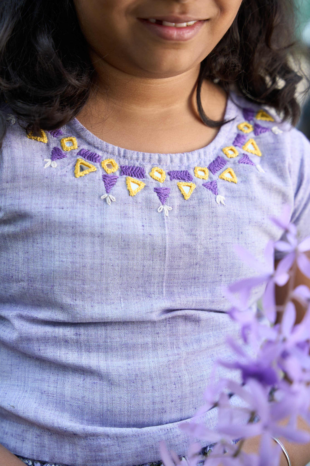 Child wearing a light blue shirt with embroidered details, holding purple flowers.