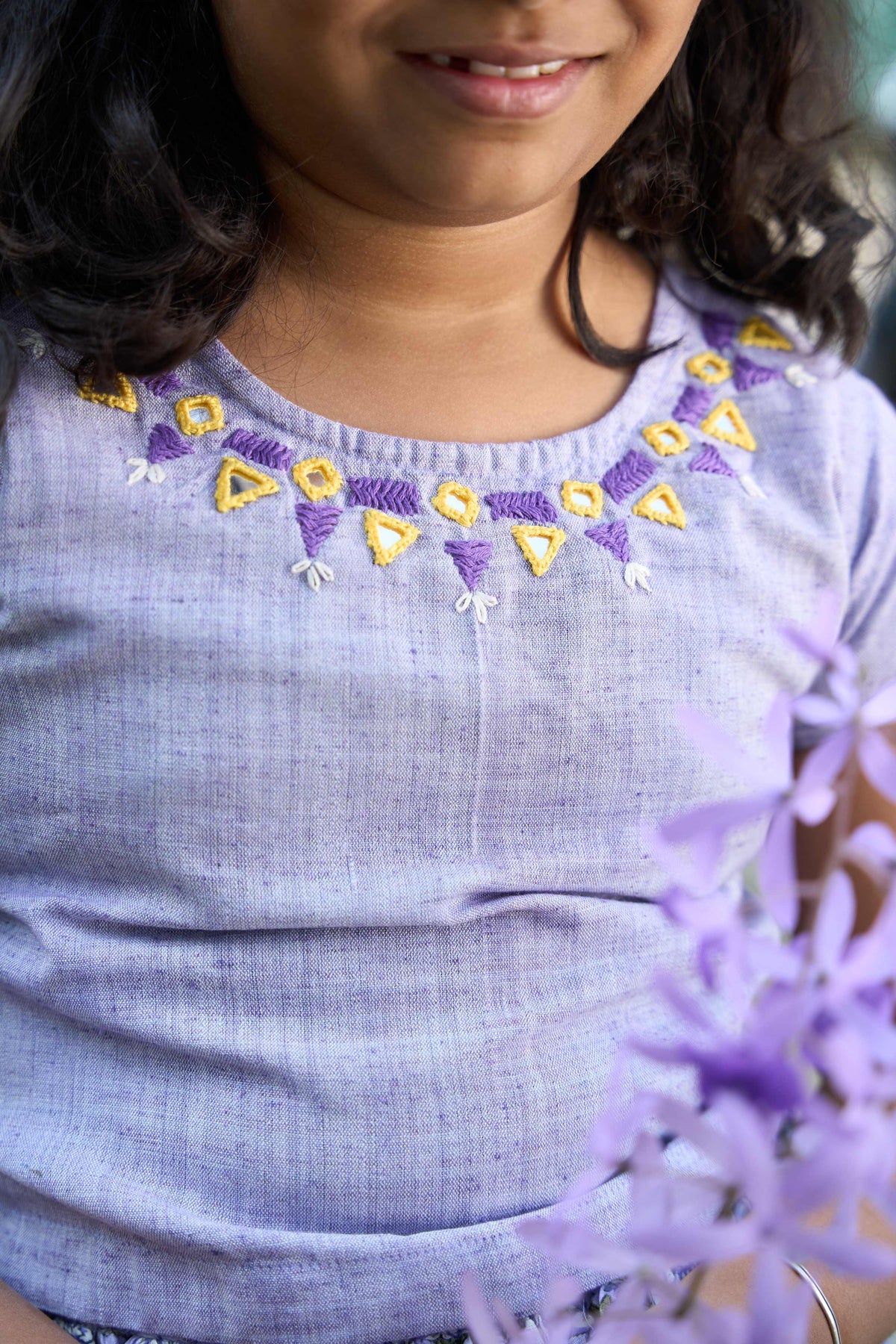 Child wearing a light blue shirt with embroidered details, holding purple flowers.