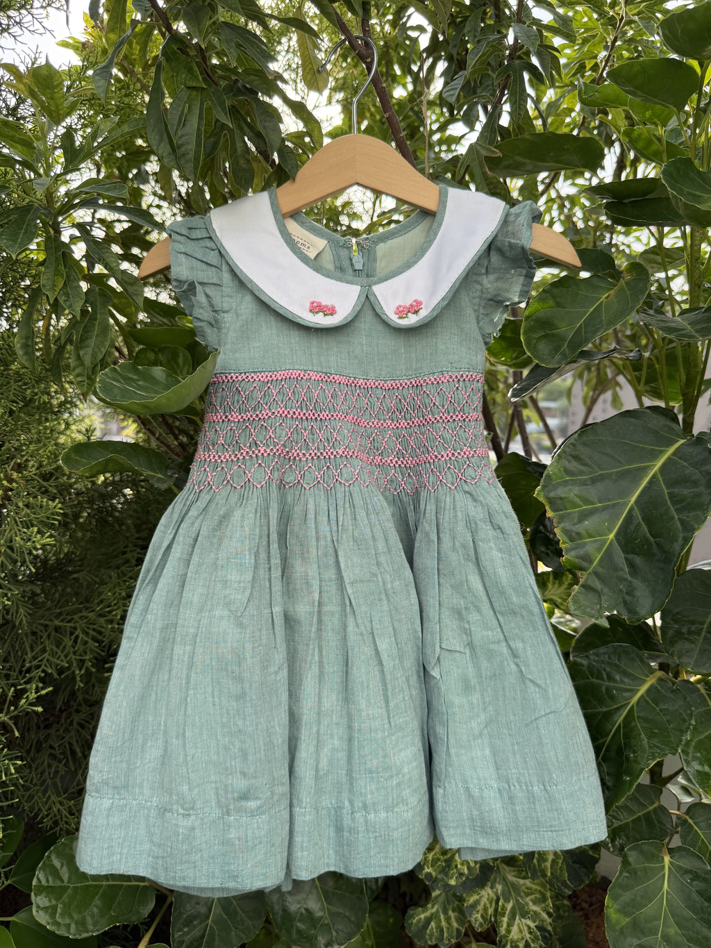 Green dress with pink embroidery on a hanger against a leafy green background
