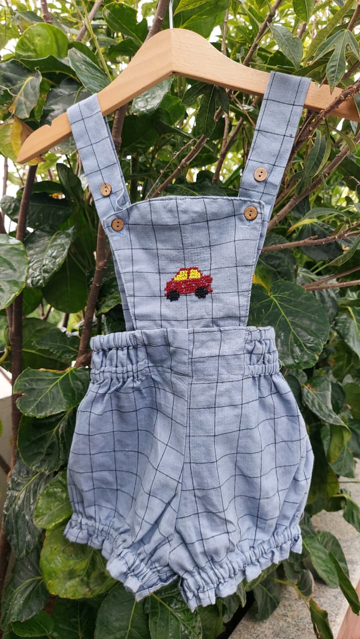 A baby romper in a light blue and white grid pattern, hanging on a wooden hanger against a background of green leaves. The romper features an embroidered red and yellow truck on the chest, decorative buttons on the shoulder straps, and a ruffled hem.