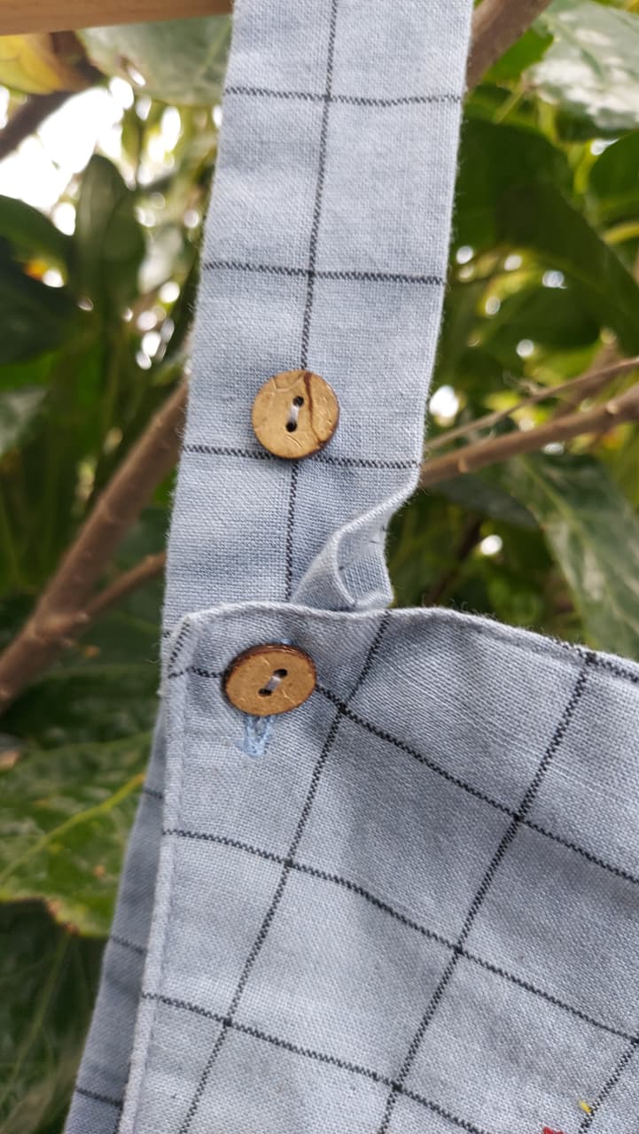 A close-up of a light blue-gray plaid fabric bag with two wooden buttons, shown outdoors against a background of green leaves and branches.