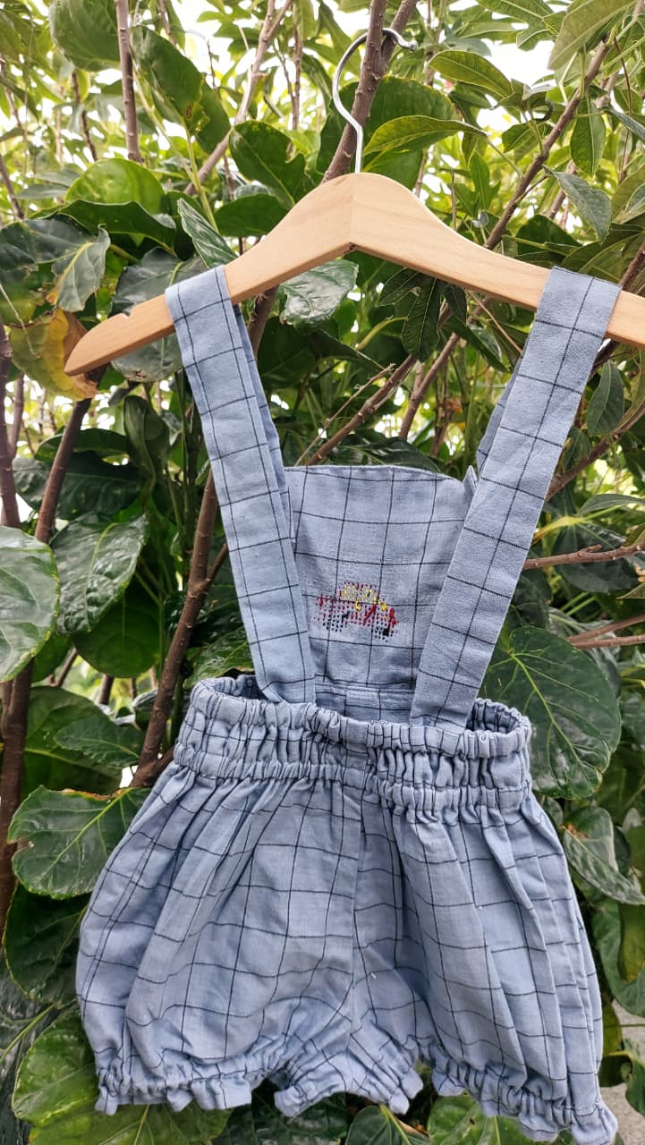 A baby romper in a light blue and white grid pattern, hanging on a wooden hanger against a background of green leaves and branches.
