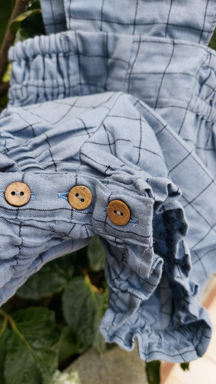 Close-up of a light blue plaid fabric garment with gathered ruffles and three wooden buttons, photographed outdoors against green leaves.