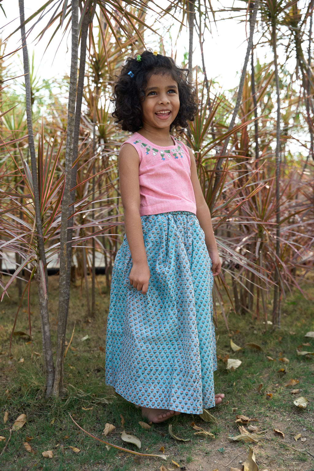 Young girl in a pink mirror work top and blue handblock print skirt standing among dried plants.