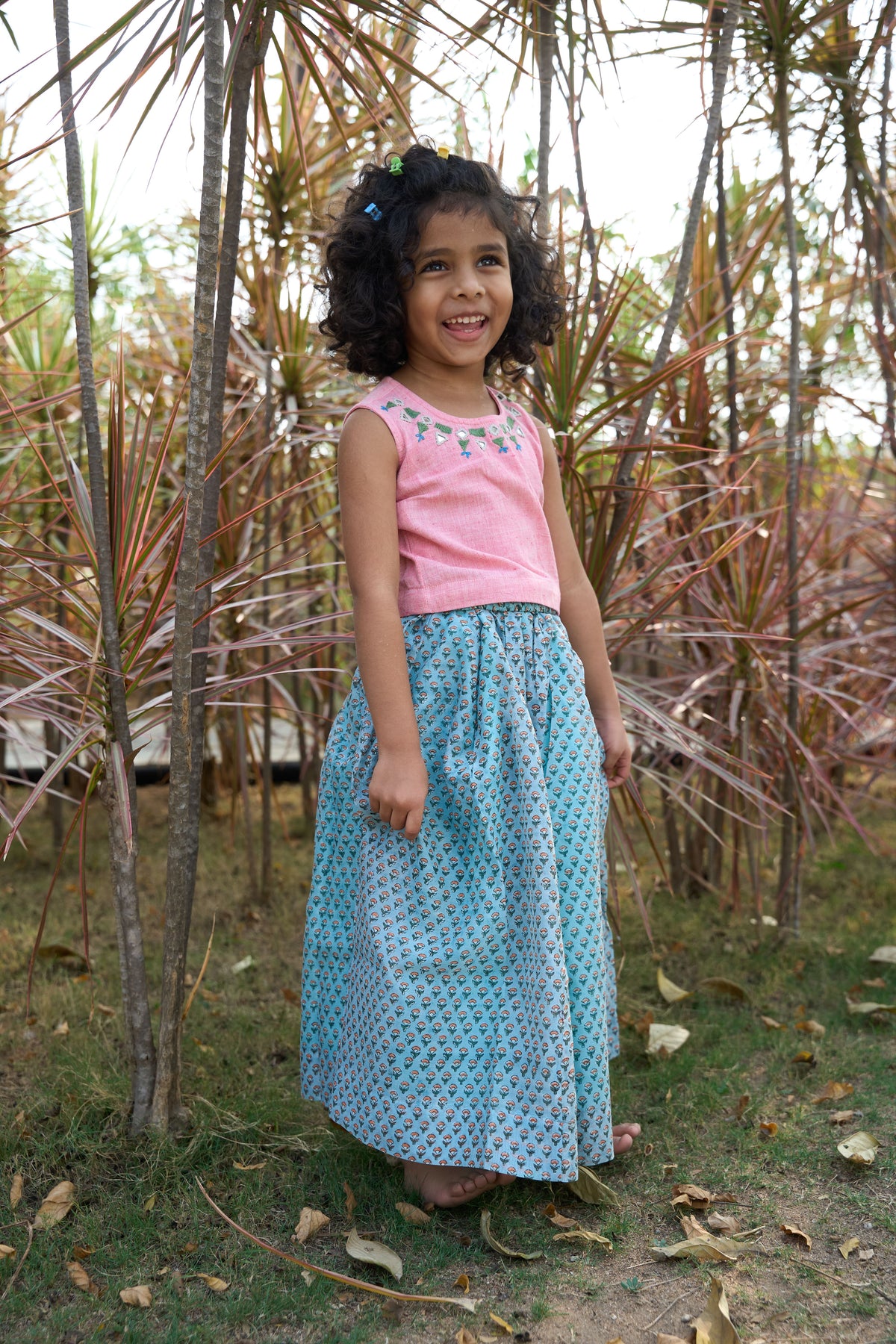 Young girl in a pink mirror work top and blue handblock print skirt standing among dried plants.