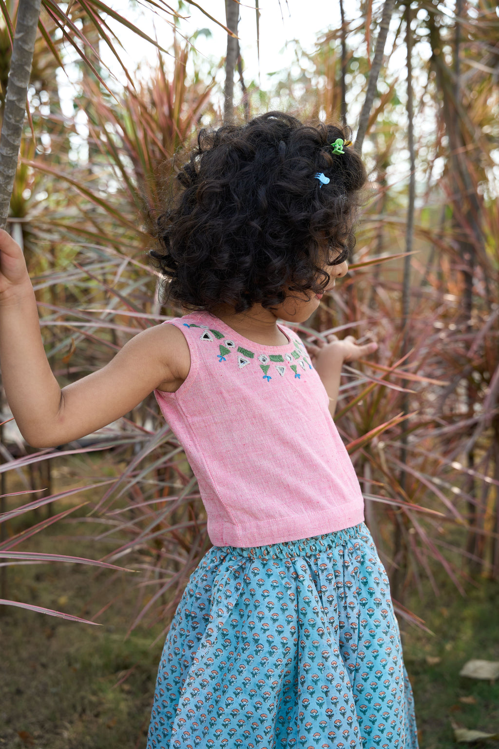 Child wearing a pink top and blue skirt standing in a natural setting with plants.