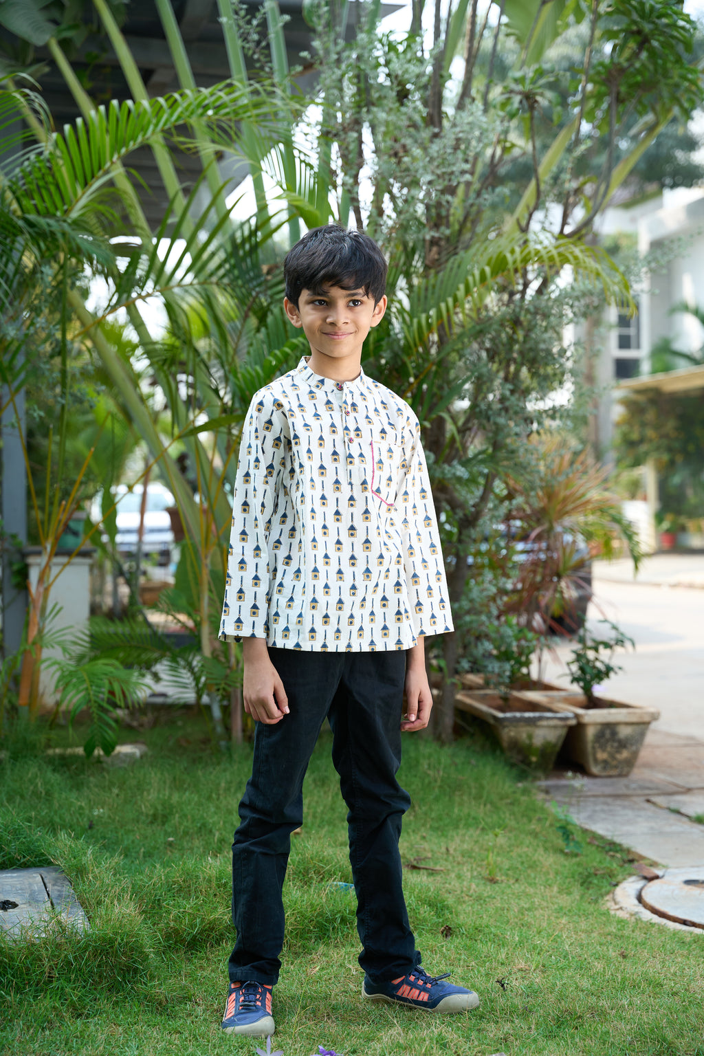 Child wearing a patterned shirt and dark pants standing outdoors with greenery in the background
