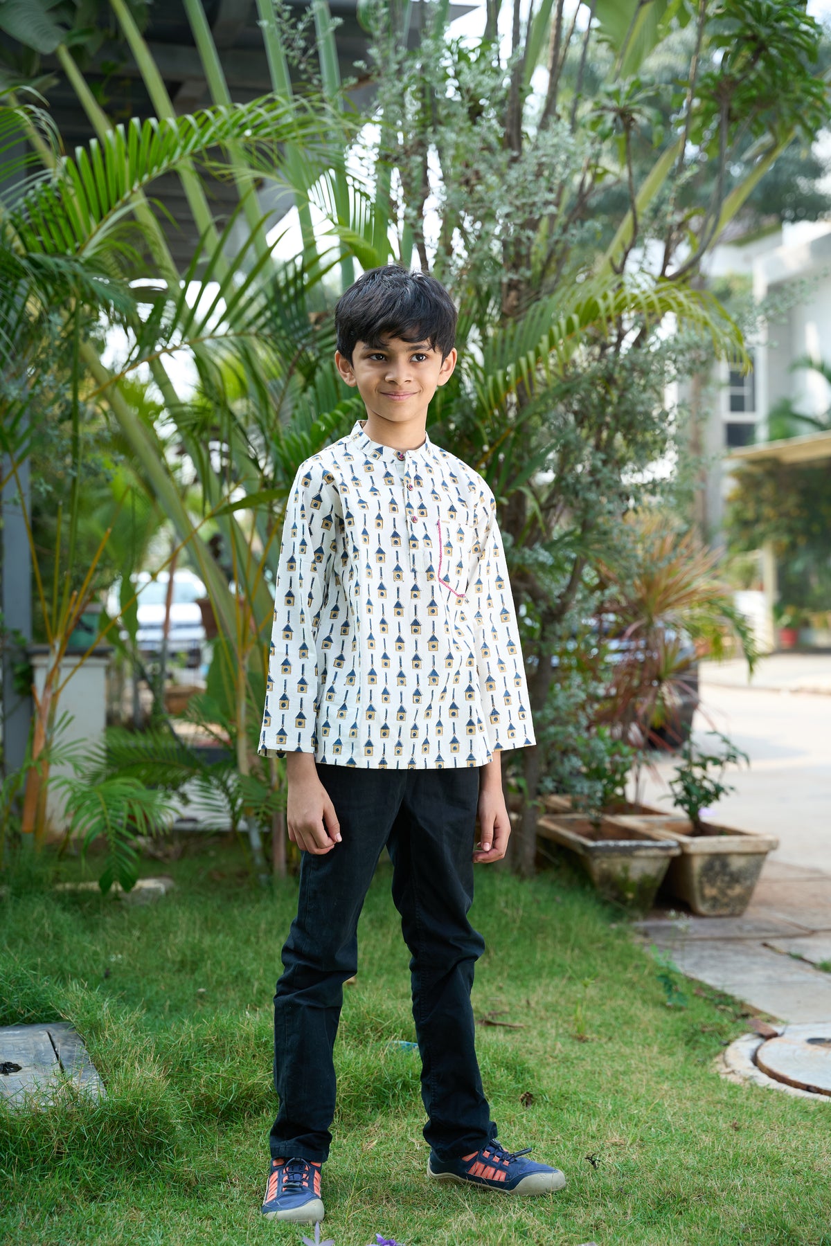 Child wearing a patterned shirt and dark pants standing outdoors with greenery in the background
