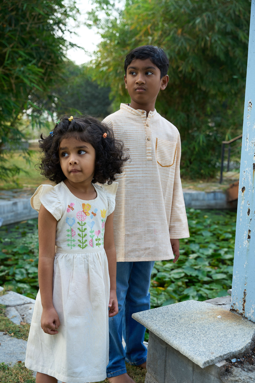 Two children standing outdoors with greenery in the background