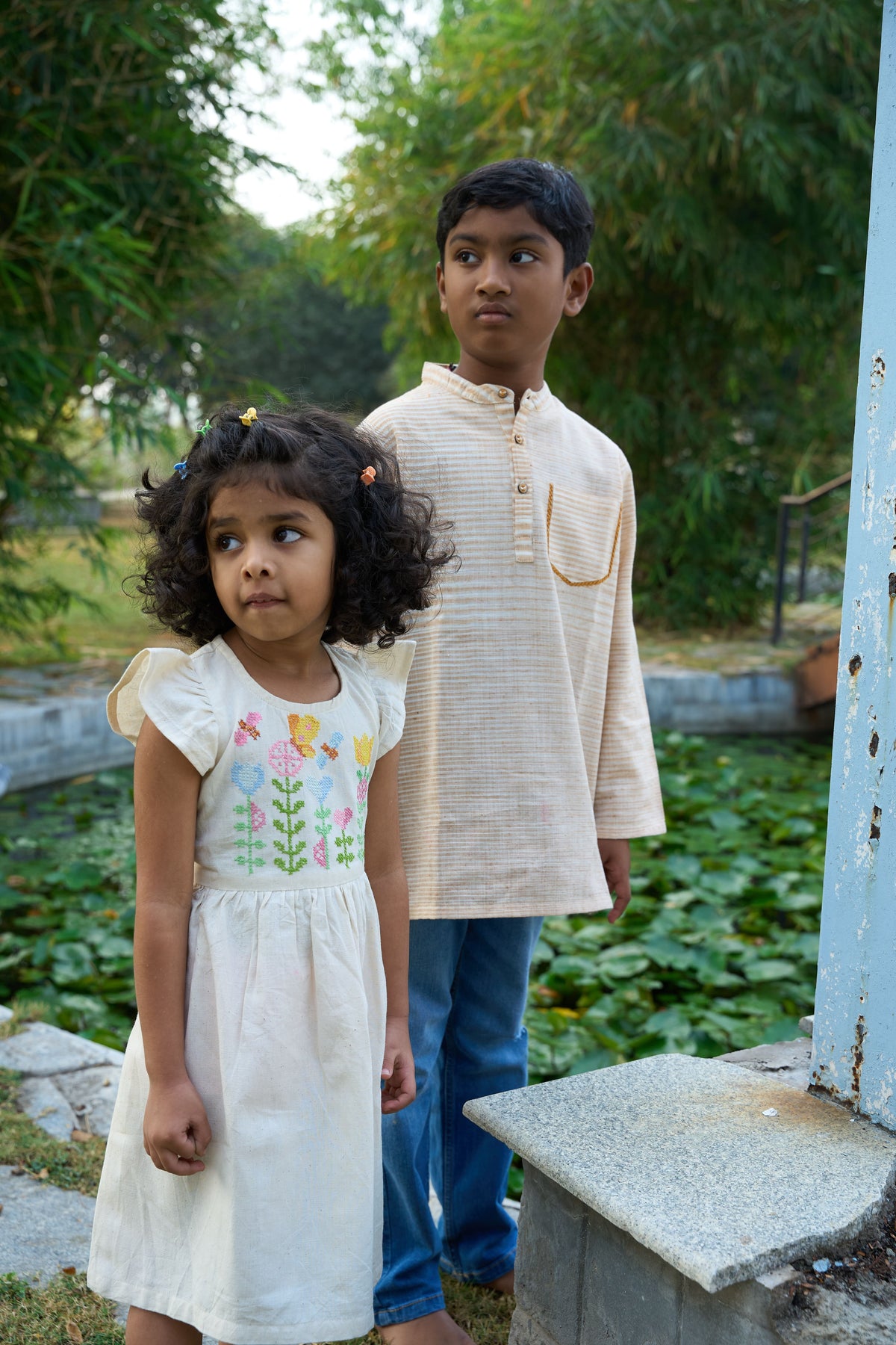 Two children standing outdoors with greenery in the background